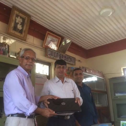 Two men exchange a laptop across a desk in a school office